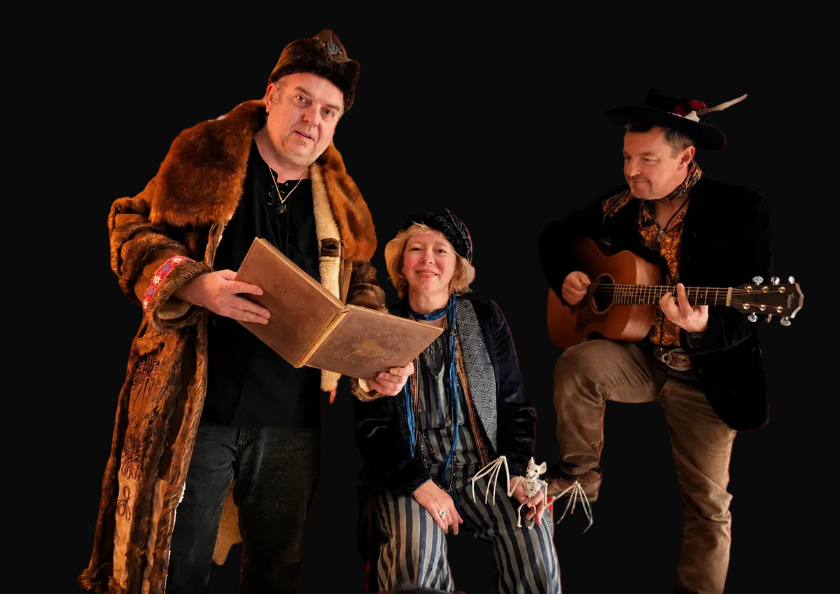 Three people in historical costume perform with an acoustic guitar. One man holds an old book, and the scene is warmly lit.
